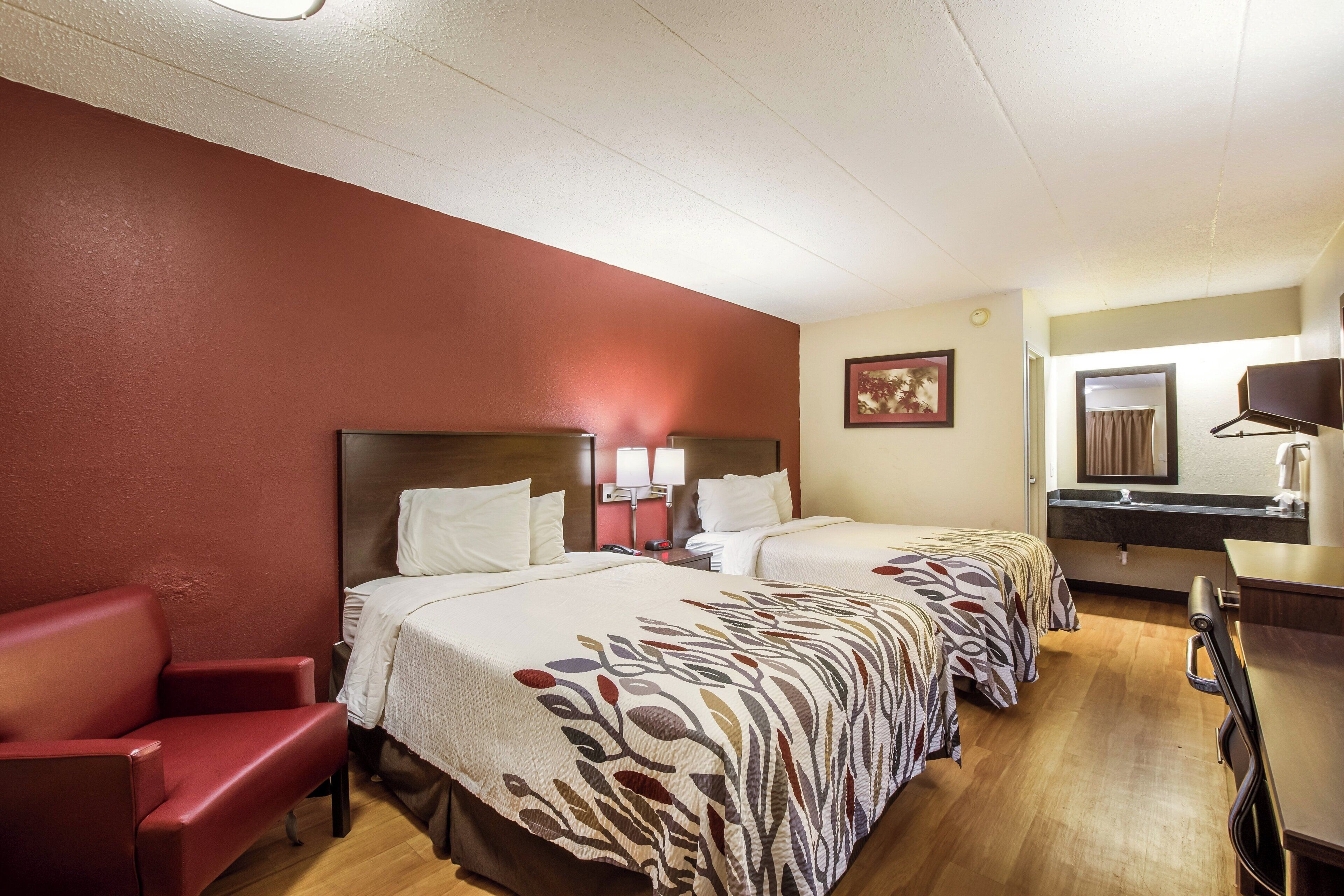 A hotel room with two beds, a red armchair, a desk with a mirror, and a small bathroom.