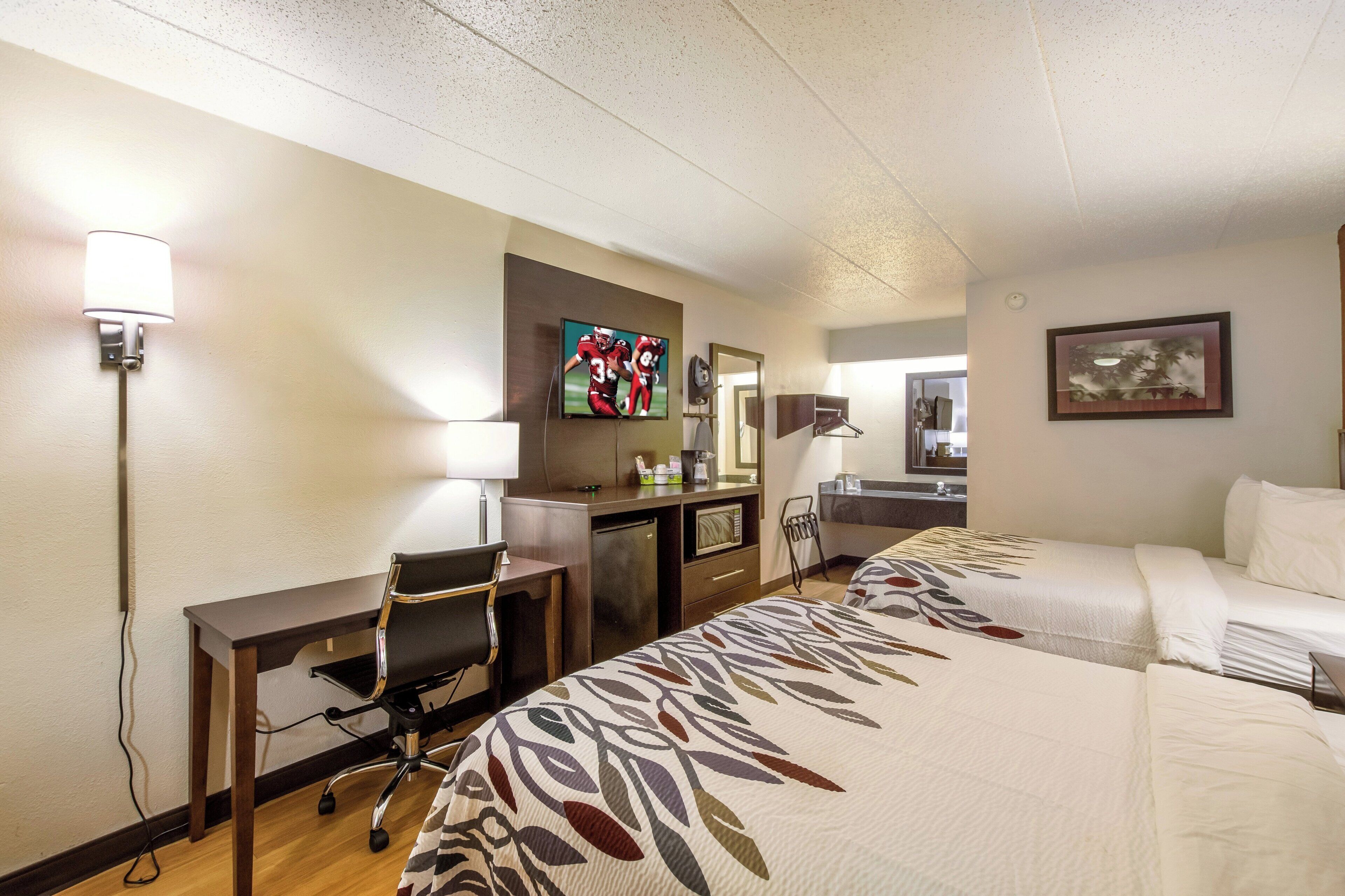 A hotel room with a desk, two beds, a TV, a mirror, and a painting.