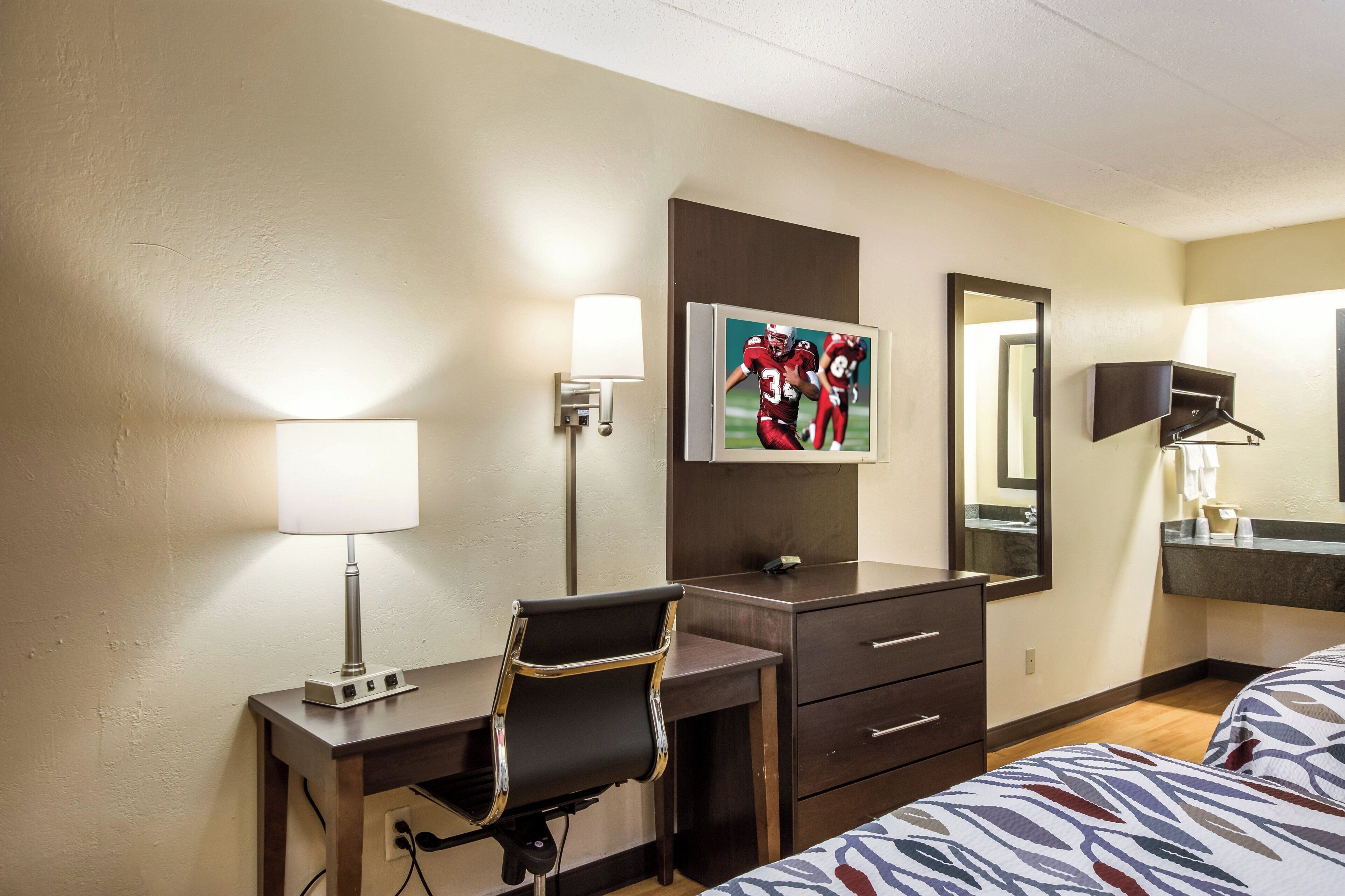 A hotel room with a desk, chair, bed, bedside table, lamp, artwork, and a mirror.