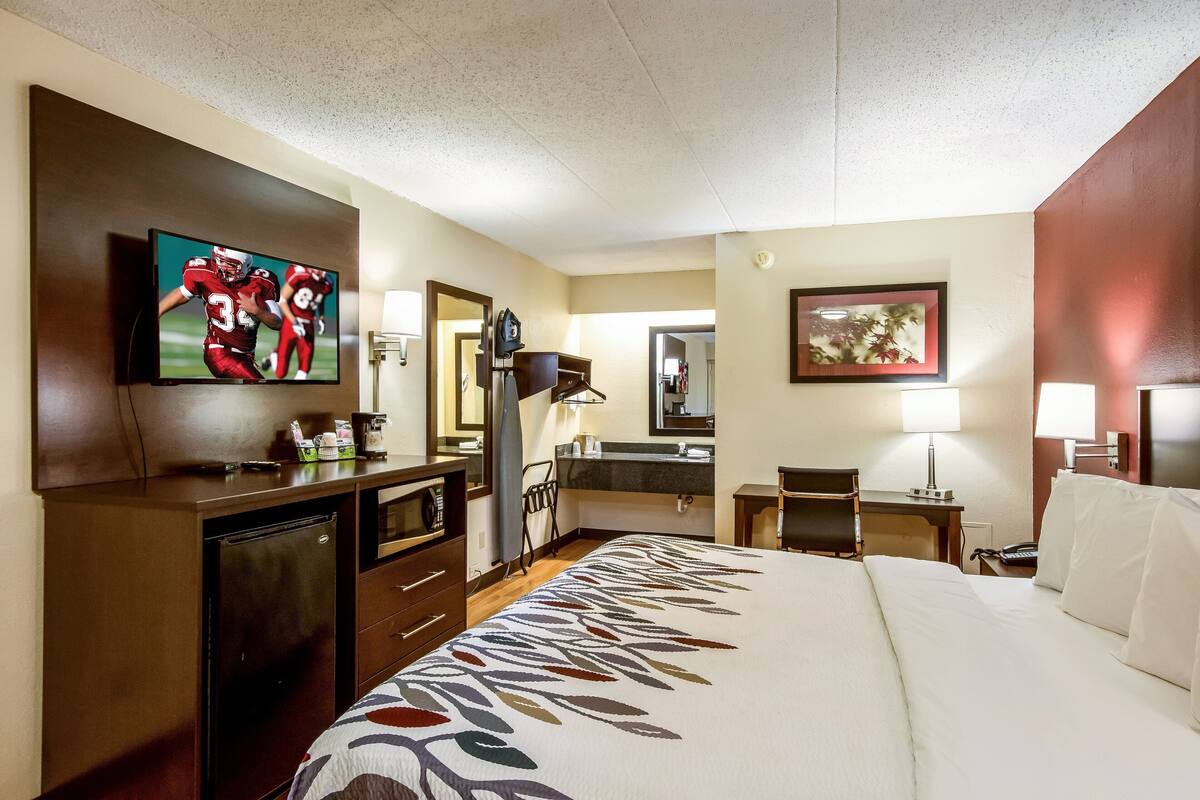 A hotel room with a large bed, a television mounted on the wall, a desk with a chair, a mirror, and a small table with a lamp.