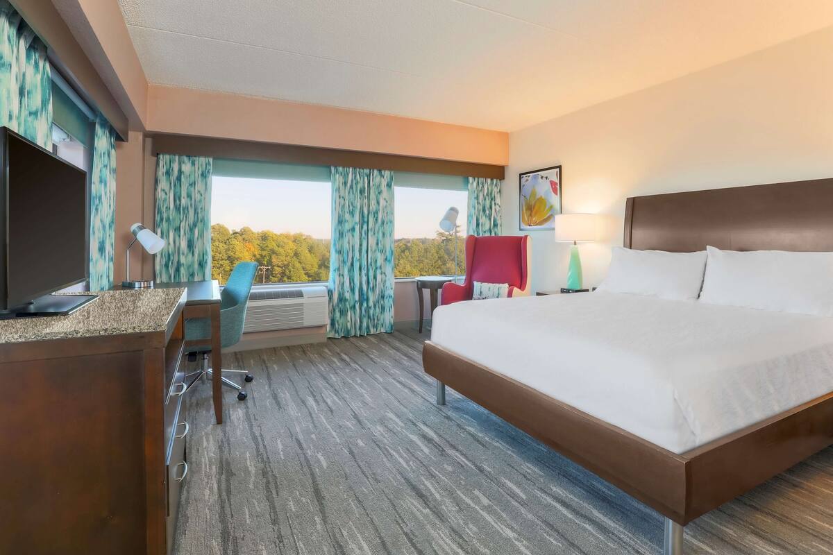 A hotel room with a bed, desk, chair, and a view of trees through the window.