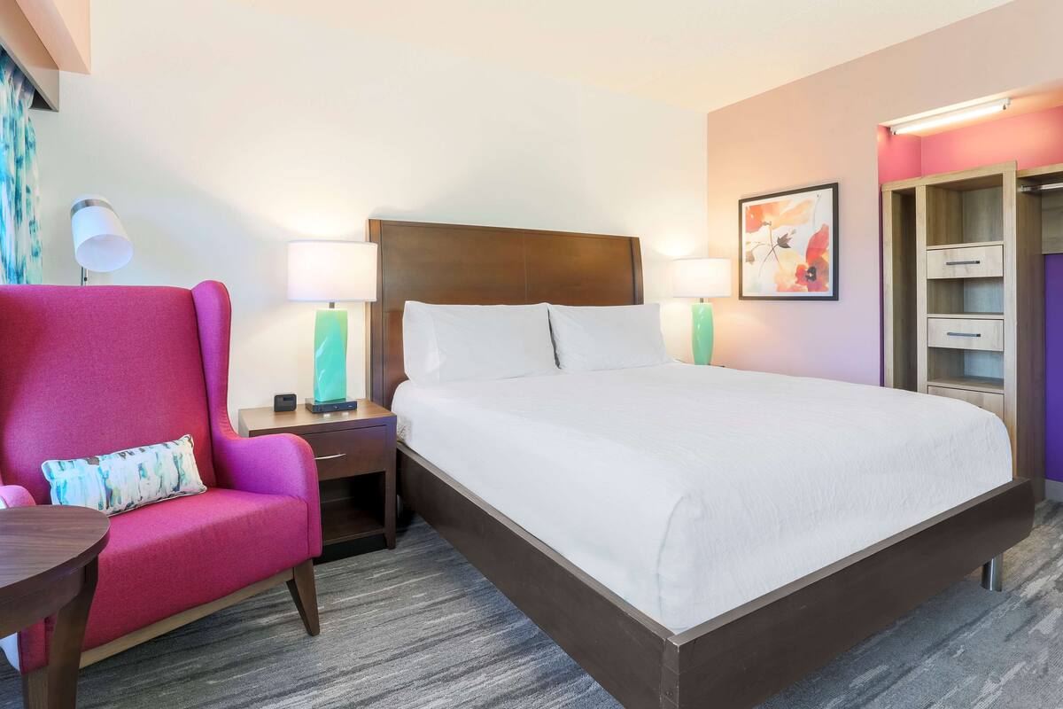 A hotel room with a large bed, a pink armchair, two bedside tables with lamps, a wooden wardrobe, and a framed picture on the wall.