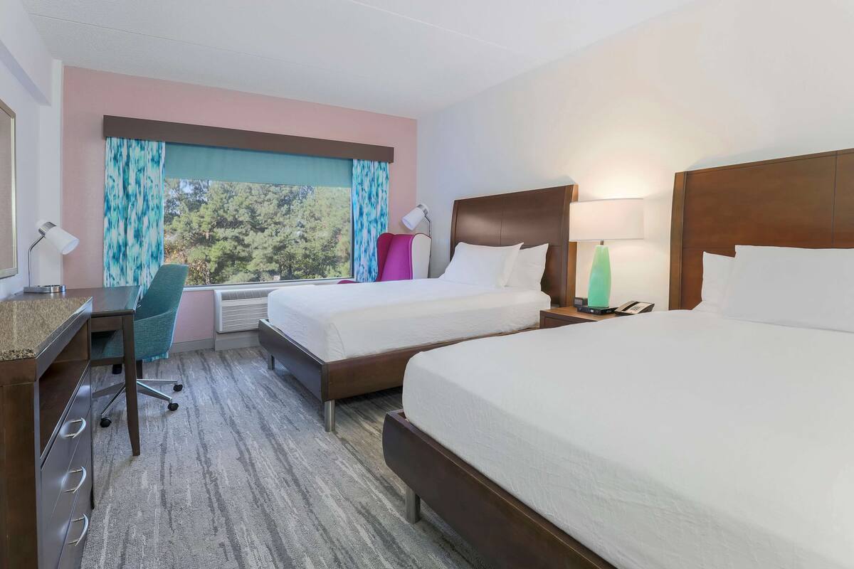 A hotel room with two beds, a desk, a chair, a lamp, and a view of trees through the window.