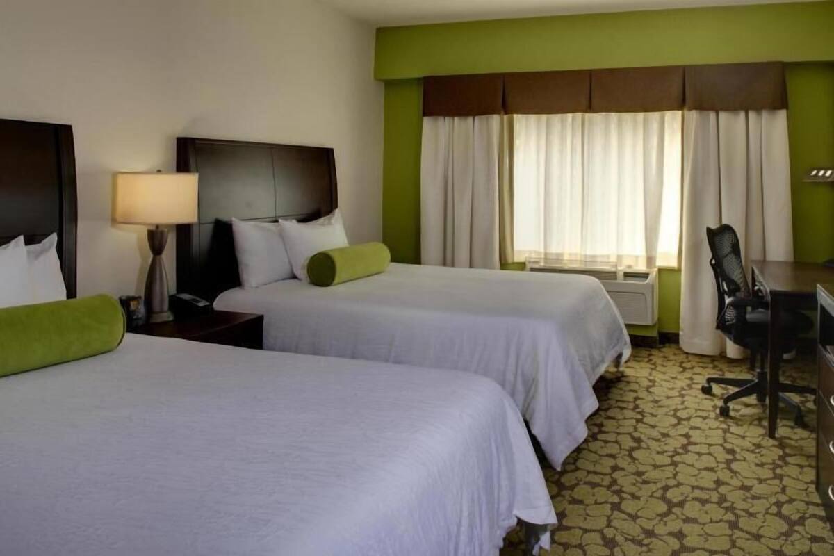 A hotel room with two beds, a desk, a chair, a lamp, and a window with curtains.