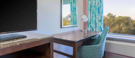 Habitación de hotel con escritorio, silla, televisor, lámpara y un ventanal con cortinas.