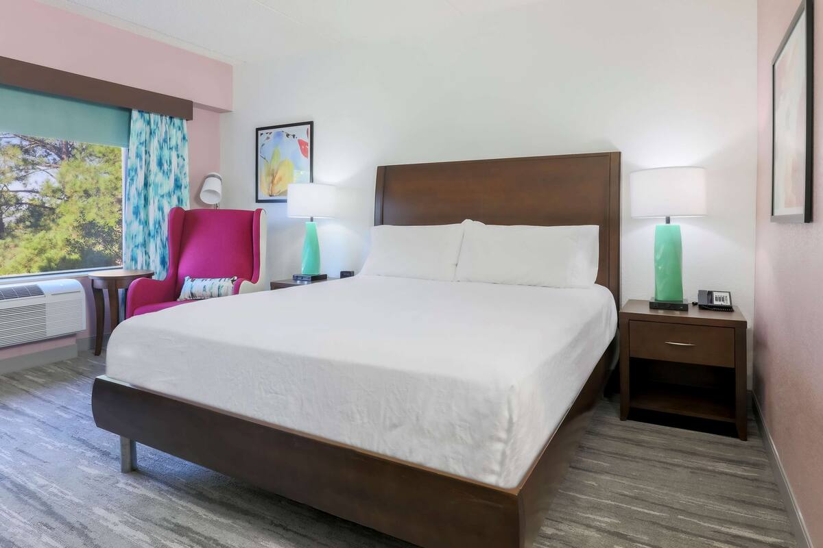 A hotel room with a large bed, two bedside tables with lamps, a chair, a window with a view of trees, and a painting on the wall.