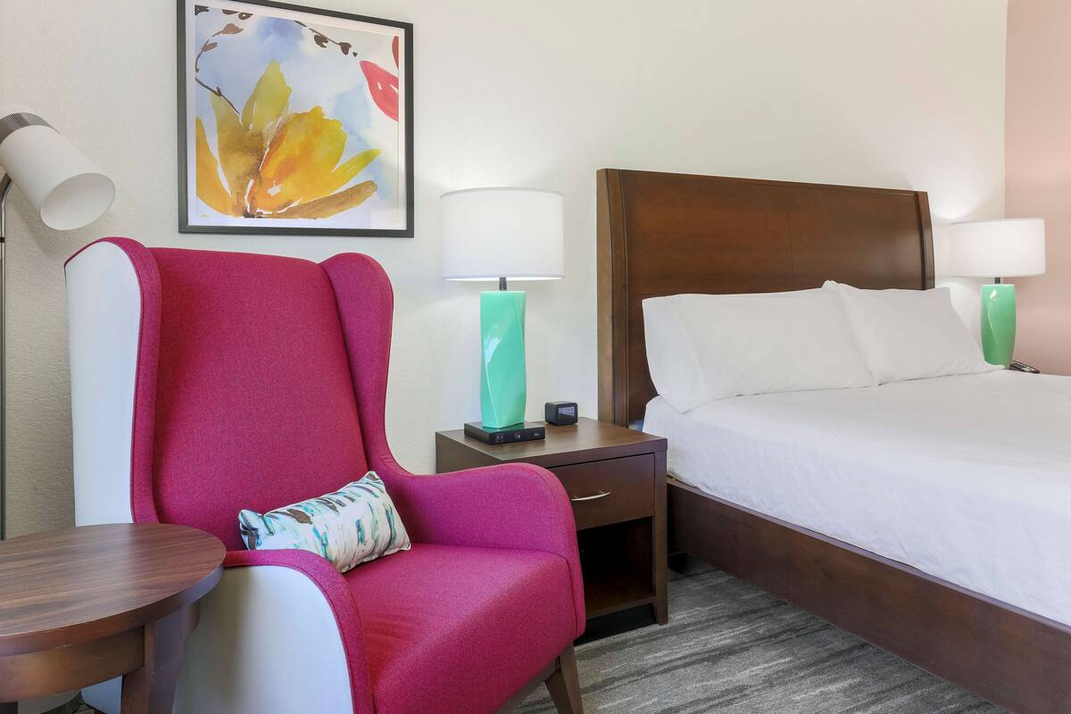 A hotel room with a bed, a nightstand with a lamp, a chair, and a table.
