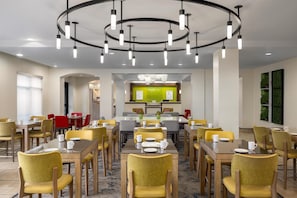 A hotel dining area with multiple tables and chairs, a central kitchen area, and a modern chandelier.