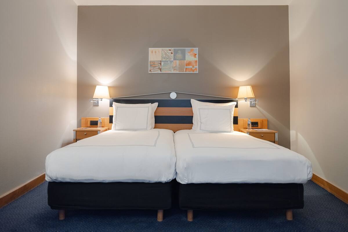 Hotellihuone, jossa on parivuode, kaksi yöpöytää, kaksi seinään kiinnitettyä lamppua ja taulu seinällä.
