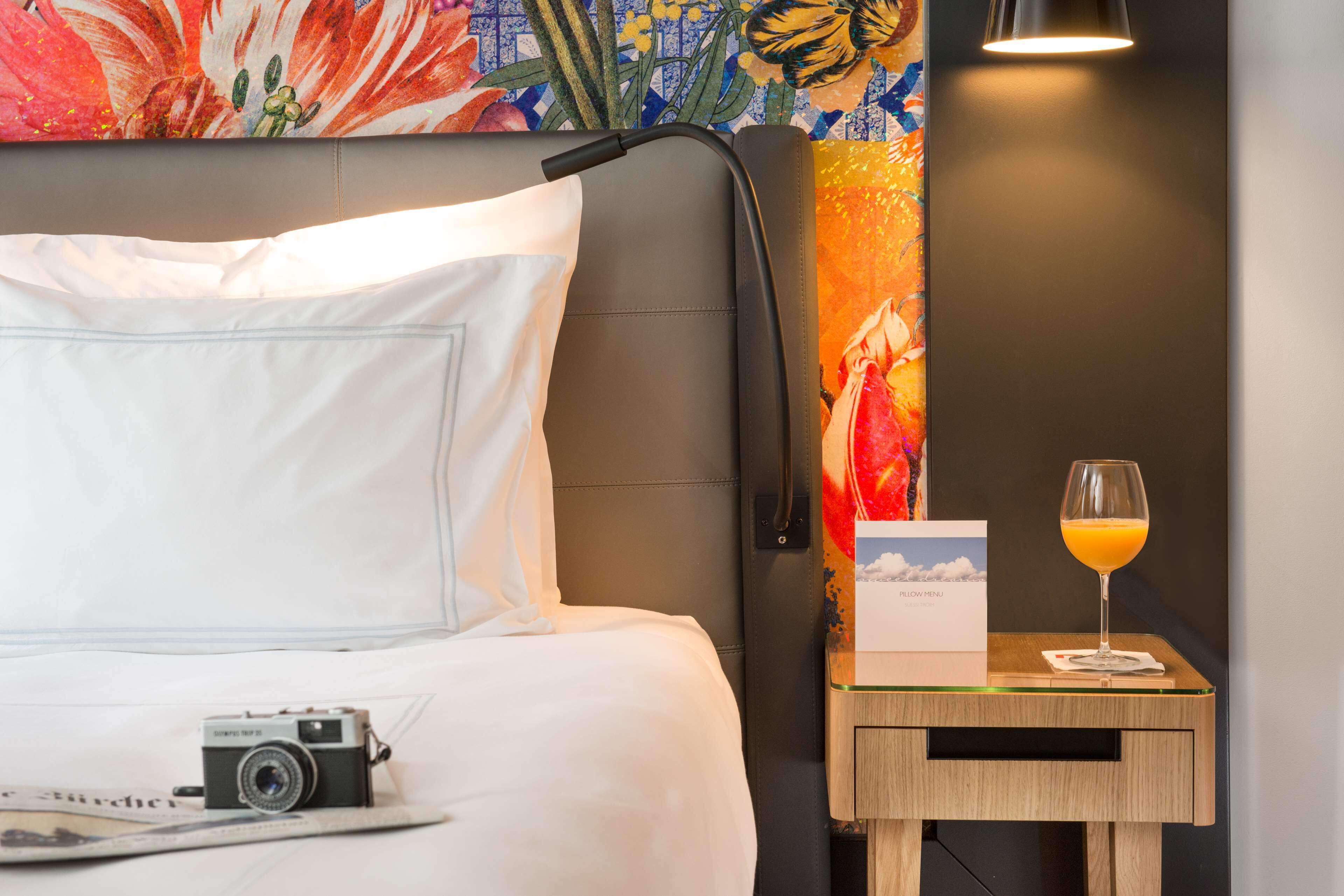 A hotel room with a bed, a nightstand with a lamp, a camera, a glass of orange juice, and a menu.