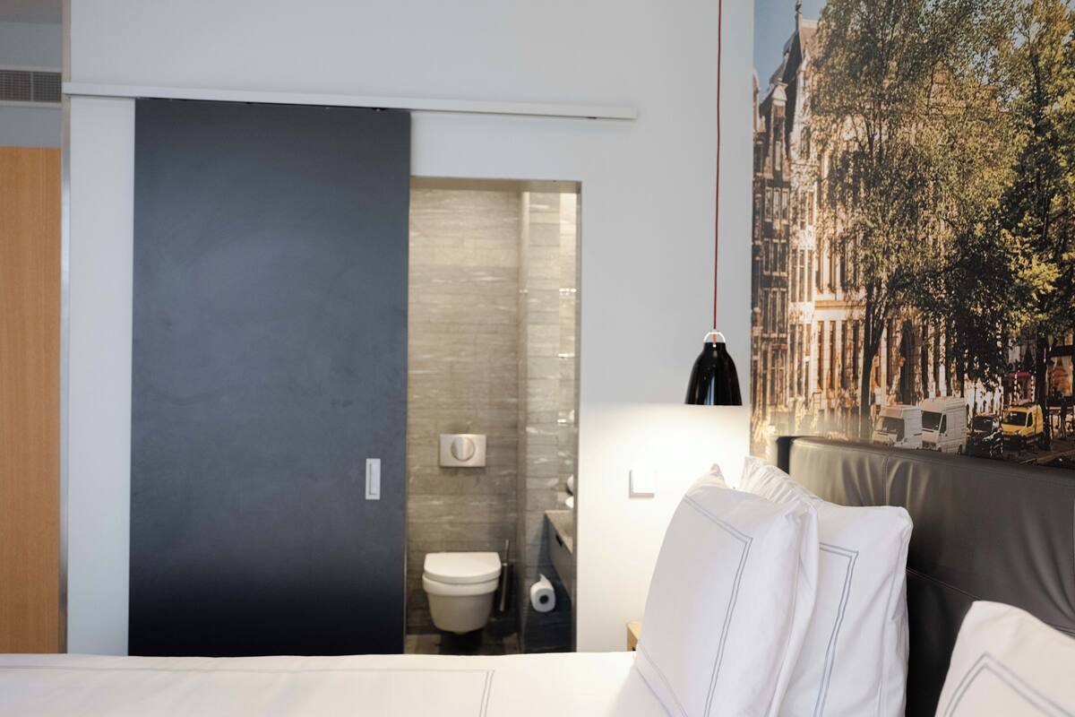 Moderni hotellihuone, jossa on sänky, valkoiset tyynyt, yöpöytä, lamppu, kylpyhuone, jossa on wc ja pesuallas, sekä seinällä oleva kaupunkimaisemaa esittävä seinämaalaus.