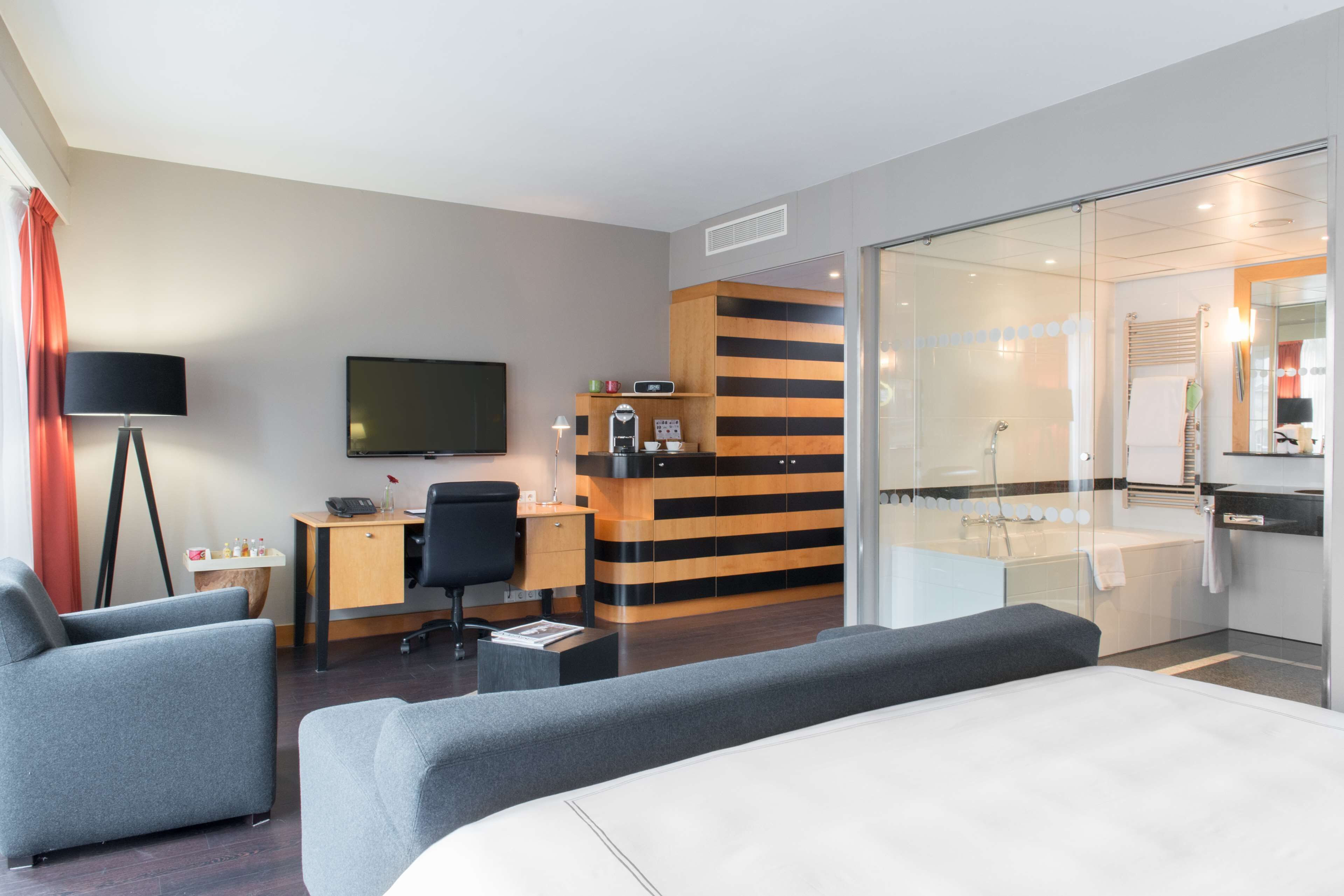 Een moderne hotelkamer met een groot bed, een bureau met stoel, een televisie, een tafeltje met lamp, en een badkamer met bad en douche.