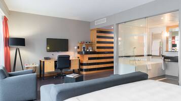 Een moderne hotelkamer met een groot bed, een bureau met stoel, een televisie, een tafeltje met lamp, en een badkamer met bad en douche.