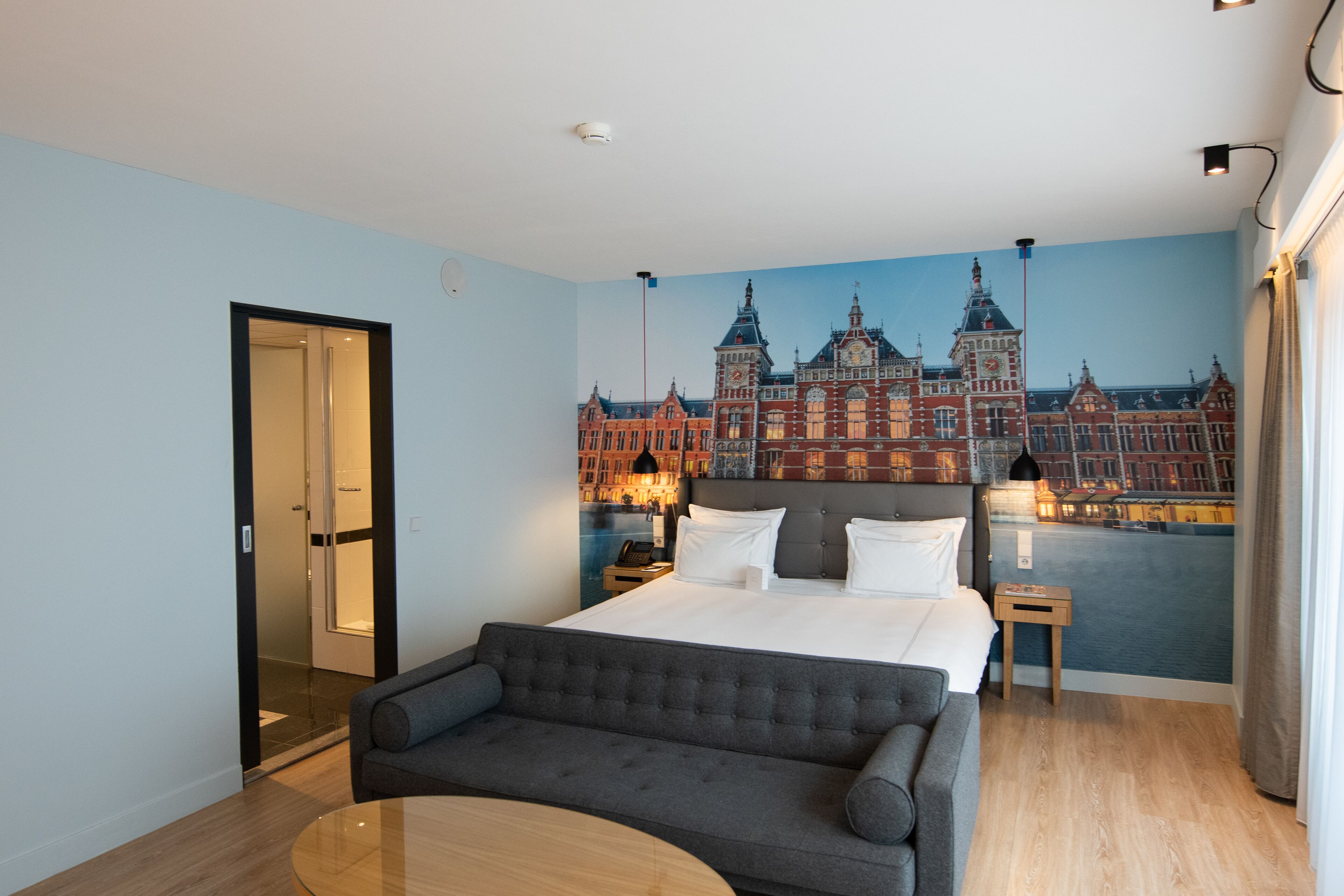 Habitación de hotel con cama, sofá, una mesita, una mesita de noche y un mural de un edificio histórico.