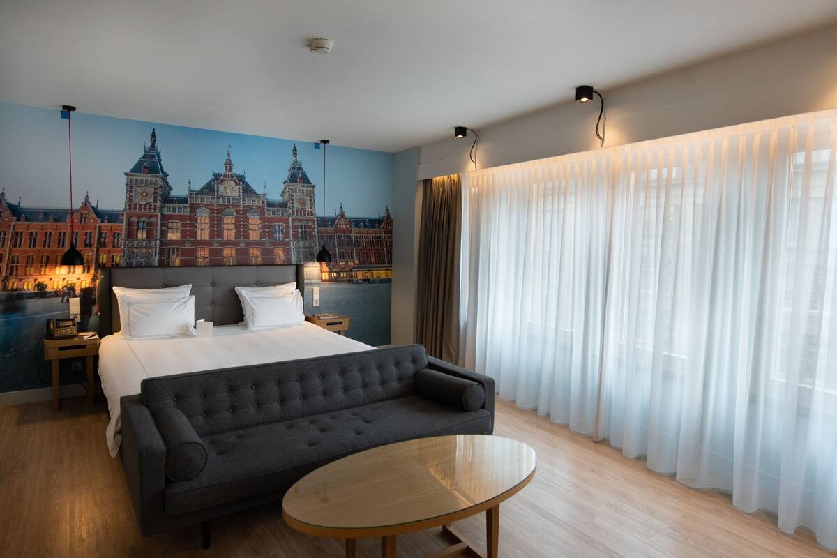 Hotellihuone, jossa on suuri sänky, sohva, pyöreä pöytä ja seinällä kaupunkimaisemaa esittävä seinämaalaus.