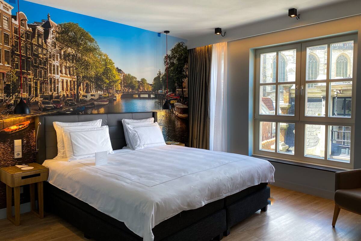 Hotellihuone, jossa on suuri sänky, yöpöytä, lamppu, ikkuna, josta avautuu näkymä rakennuksiin ja kanavalle, sekä seinämaalaus, jossa on kuvattuna kaupunkimaisema.