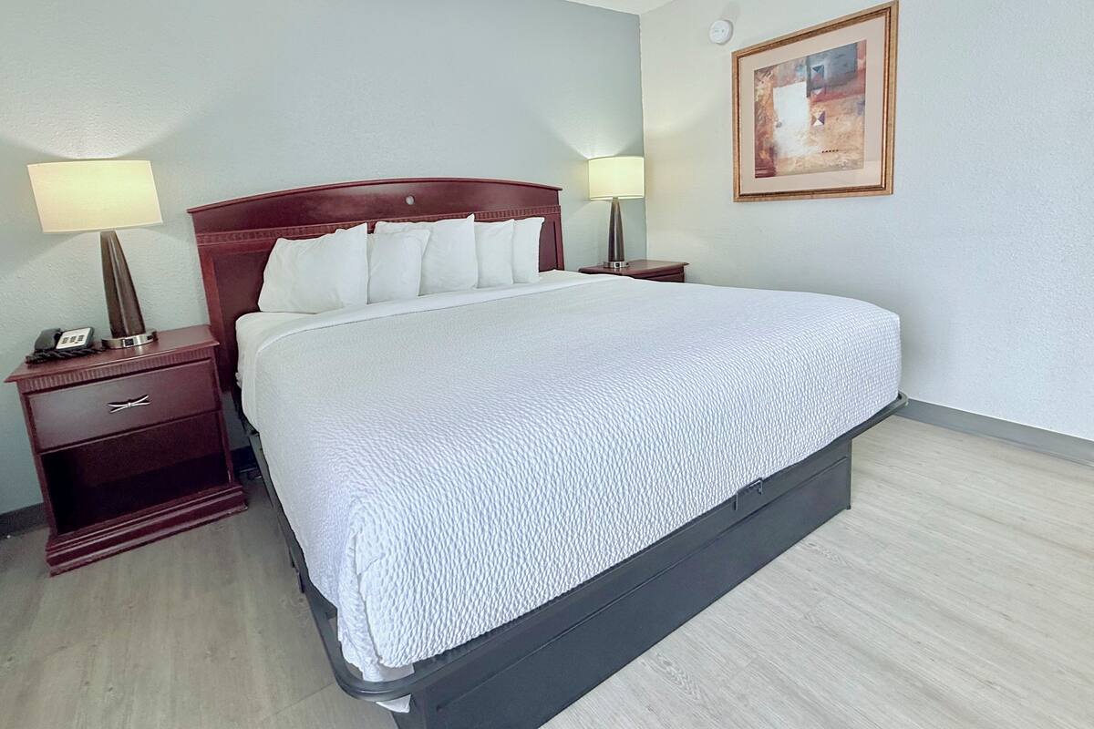 A hotel room with a large bed, two bedside tables with lamps, a nightstand, a framed picture on the wall, and a light-colored wooden floor.
