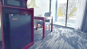 Habitación de hotel con refrigerador, lámpara, mesa y vistas al exterior.
