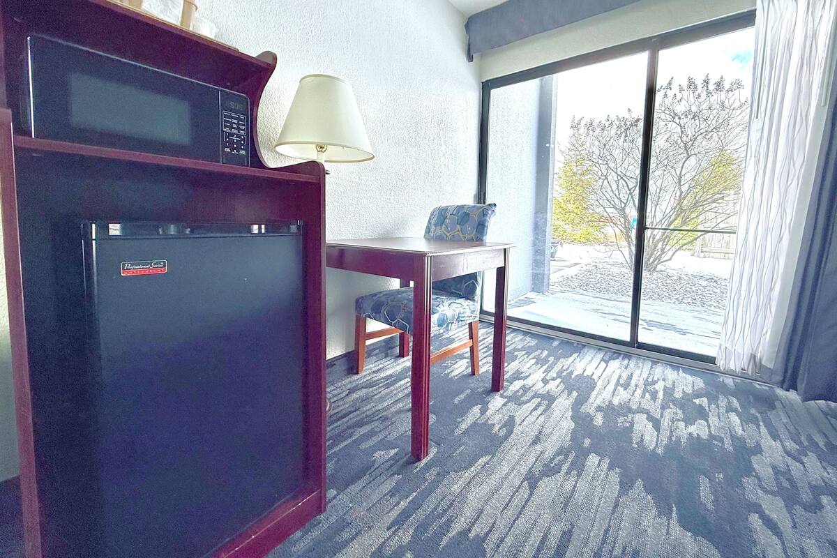 A hotel room with a refrigerator, a lamp, a table, and a view of the outdoors.