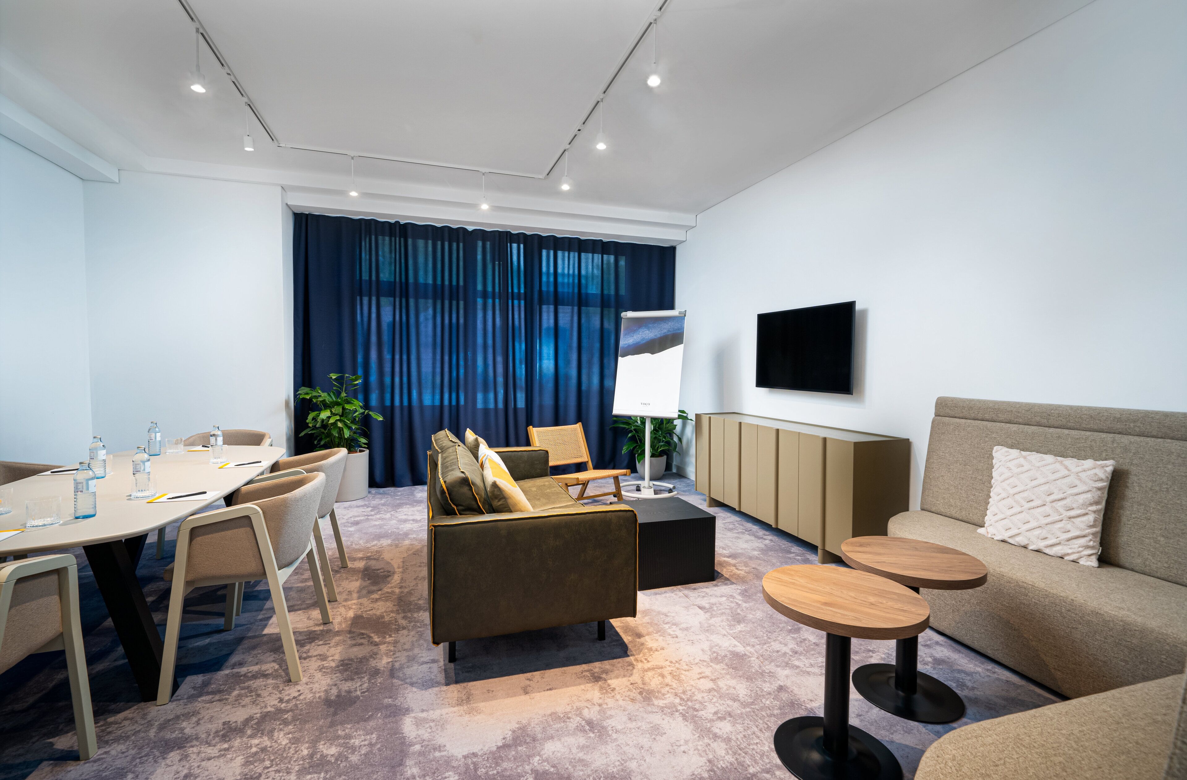A modern hotel room with a dining area, a sofa, two small tables, and a TV mounted on the wall.