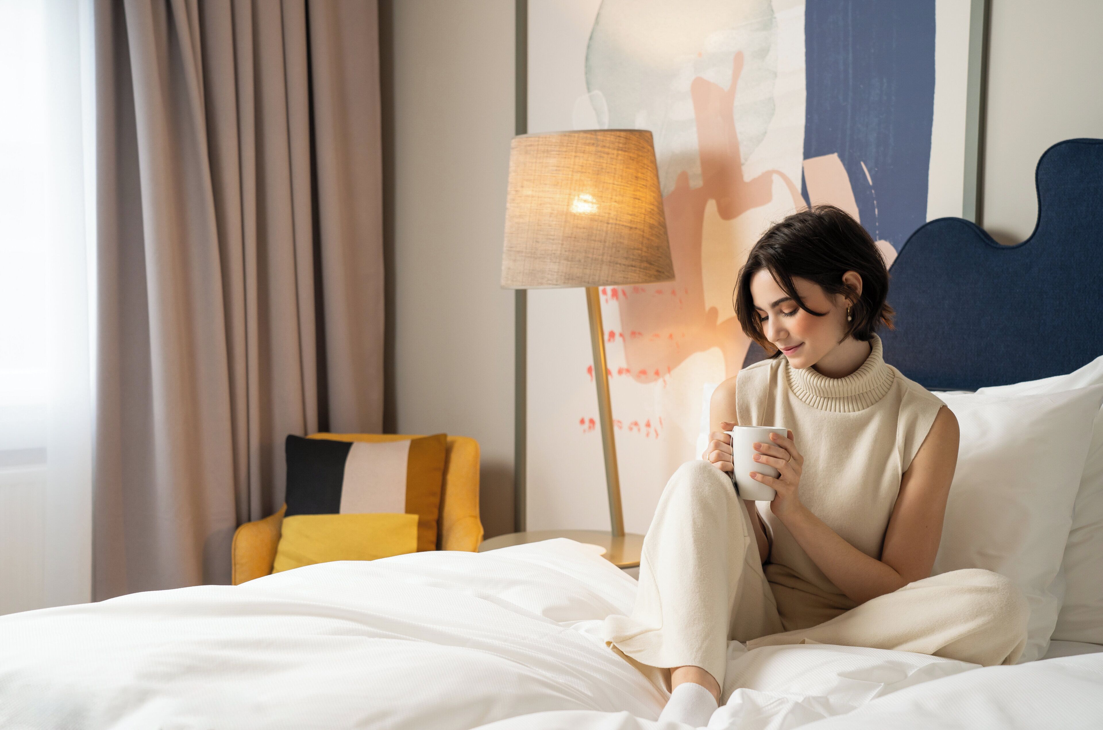 Eine Person sitzt auf einem Bett mit weißer Bettwäsche, eine Tasse haltend, in einem Raum mit einer Lampe und einem Gemälde an der Wand.