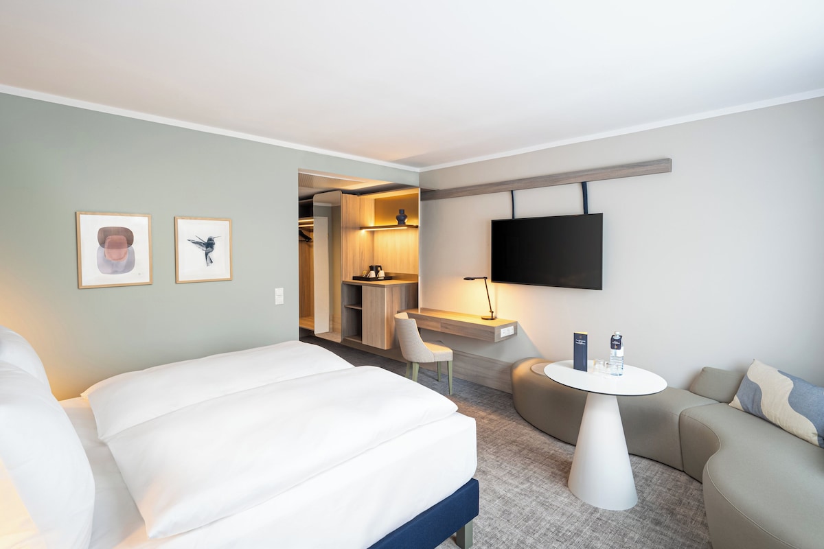 A modern hotel room with a large bed, a sofa, a small table, a TV, and a desk area with a chair.