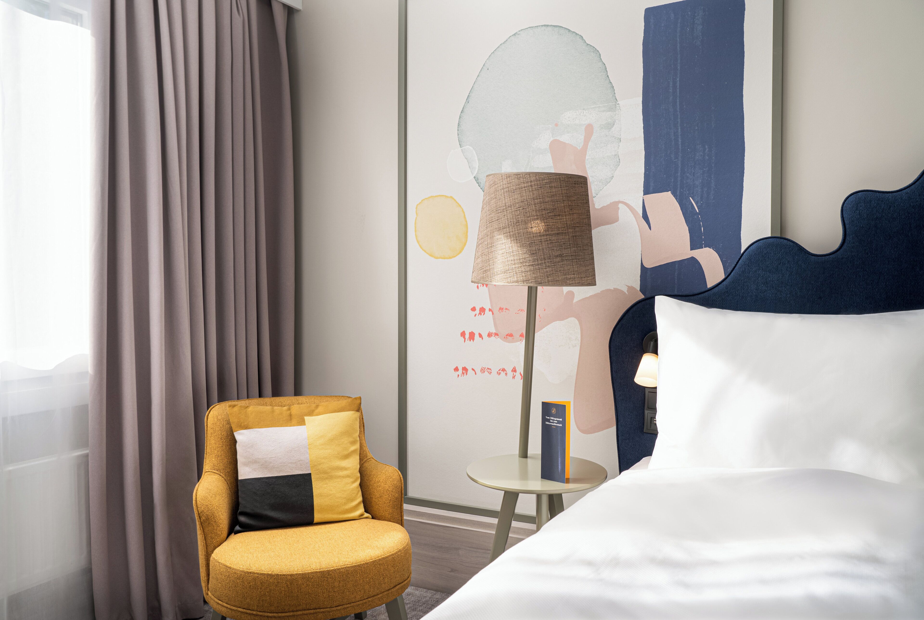 A hotel room with a bed, a round armchair with a pillow, a small table with a lamp, and a wall with abstract art.