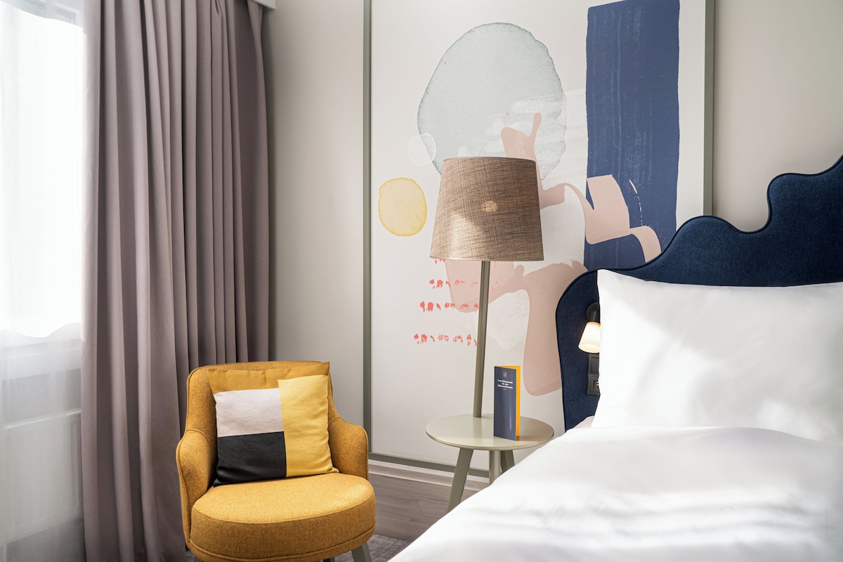 A hotel room with a bed, a chair, a lamp, and a painting on the wall.