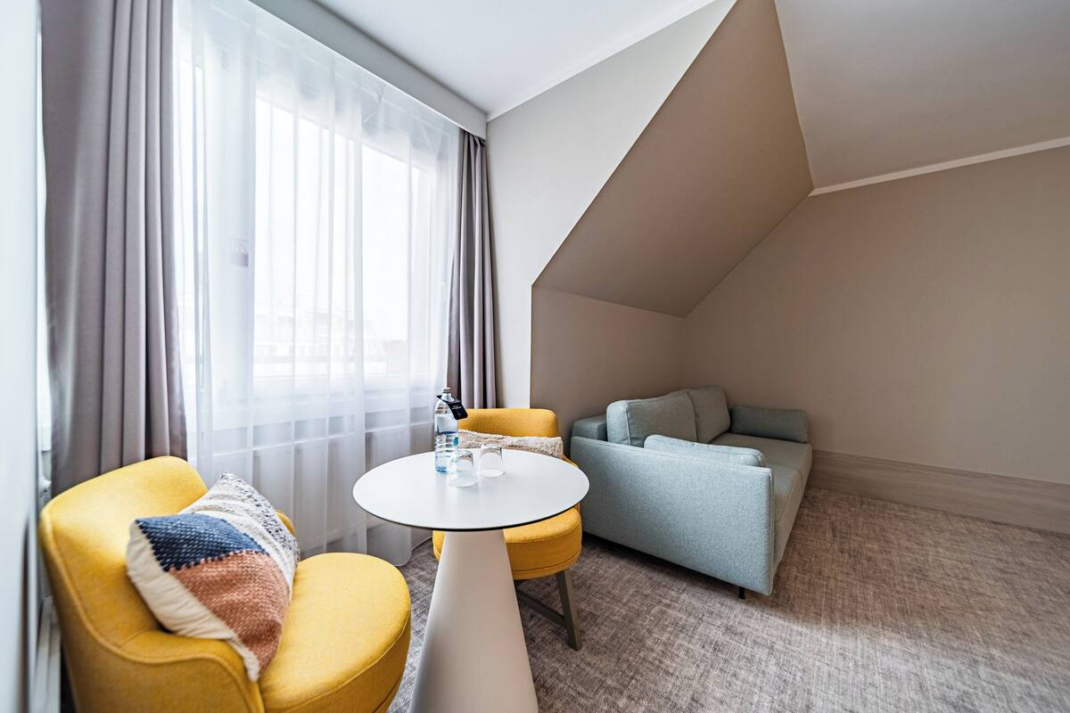 A hotel room with a yellow armchair, a gray sofa, a small round table, and a large window with sheer curtains.