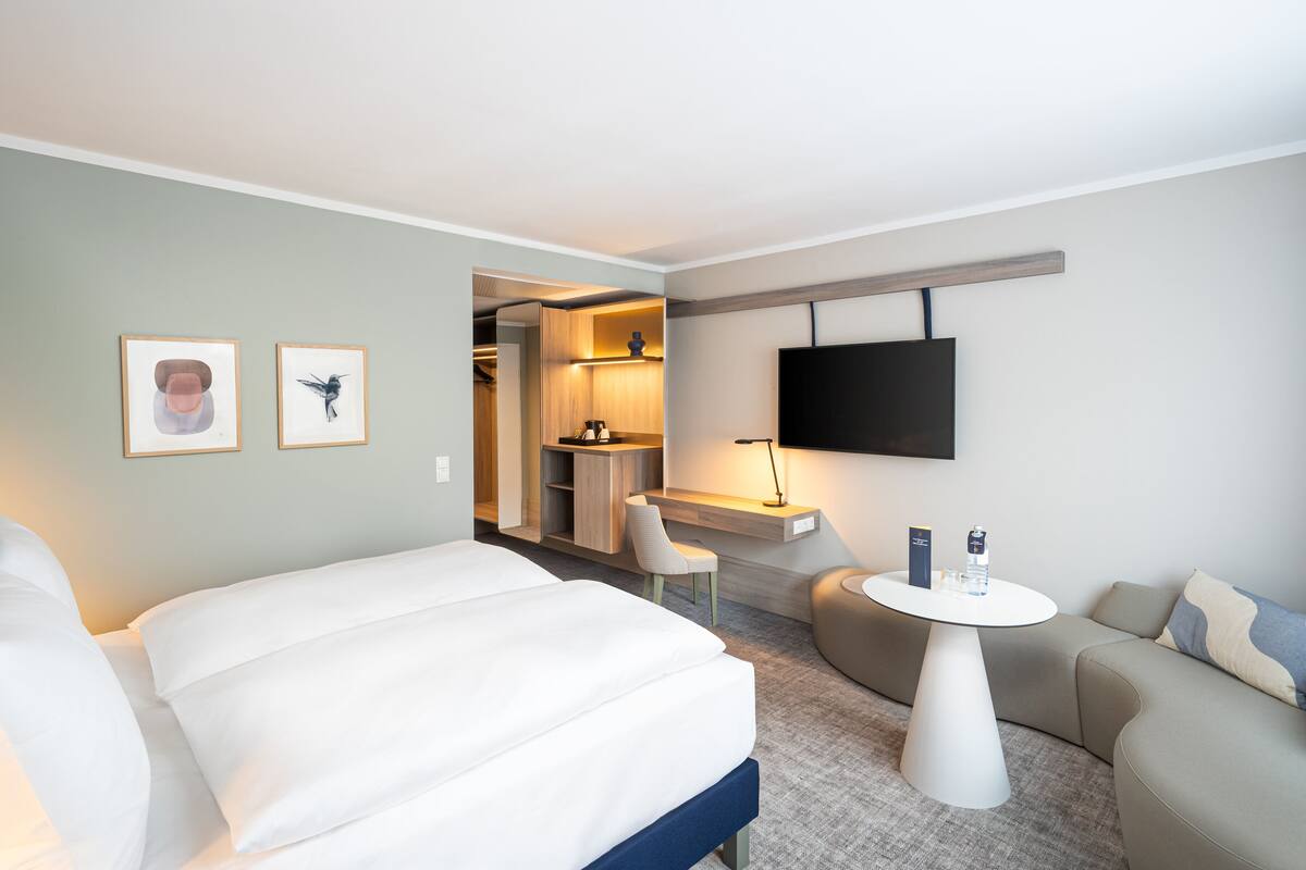 A modern hotel room with a large bed, a sofa, a small table, a TV, and a desk area with a chair.