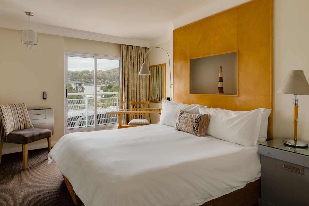 Habitación de hotel con una cama grande, una silla, un escritorio con lámpara, un espejo y vistas al exterior.