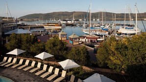 Un puerto deportivo con barcos amarrados, sillones de descanso, sombrillas y vistas a colinas al fondo.