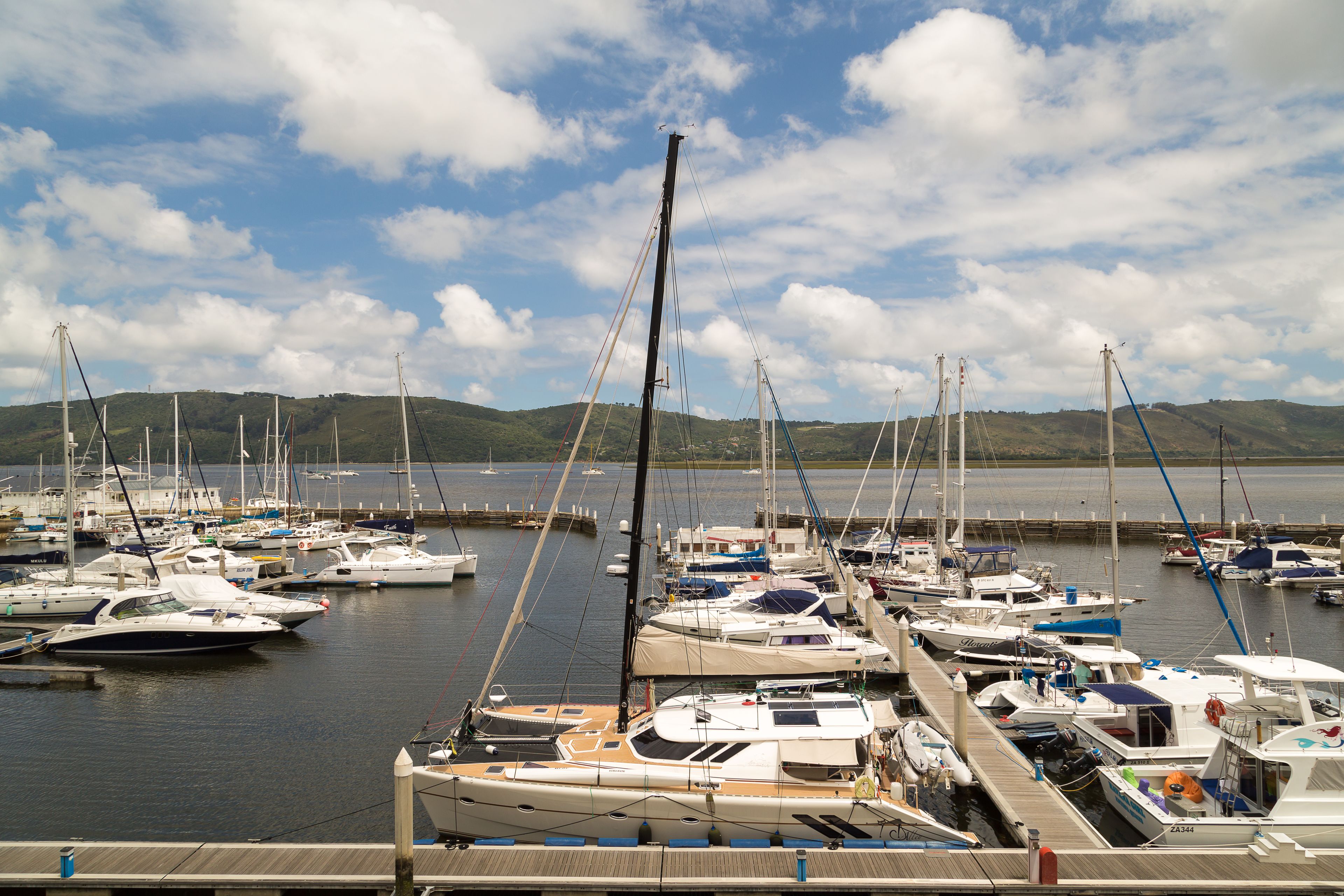 A marina with multiple boats docked, surrounded by hills and a partly cloudy sky.