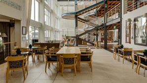 Un espacio interior amplio con varias mesas y sillas de comedor, una escalera central y ventanas grandes que permiten la entrada de luz natural.