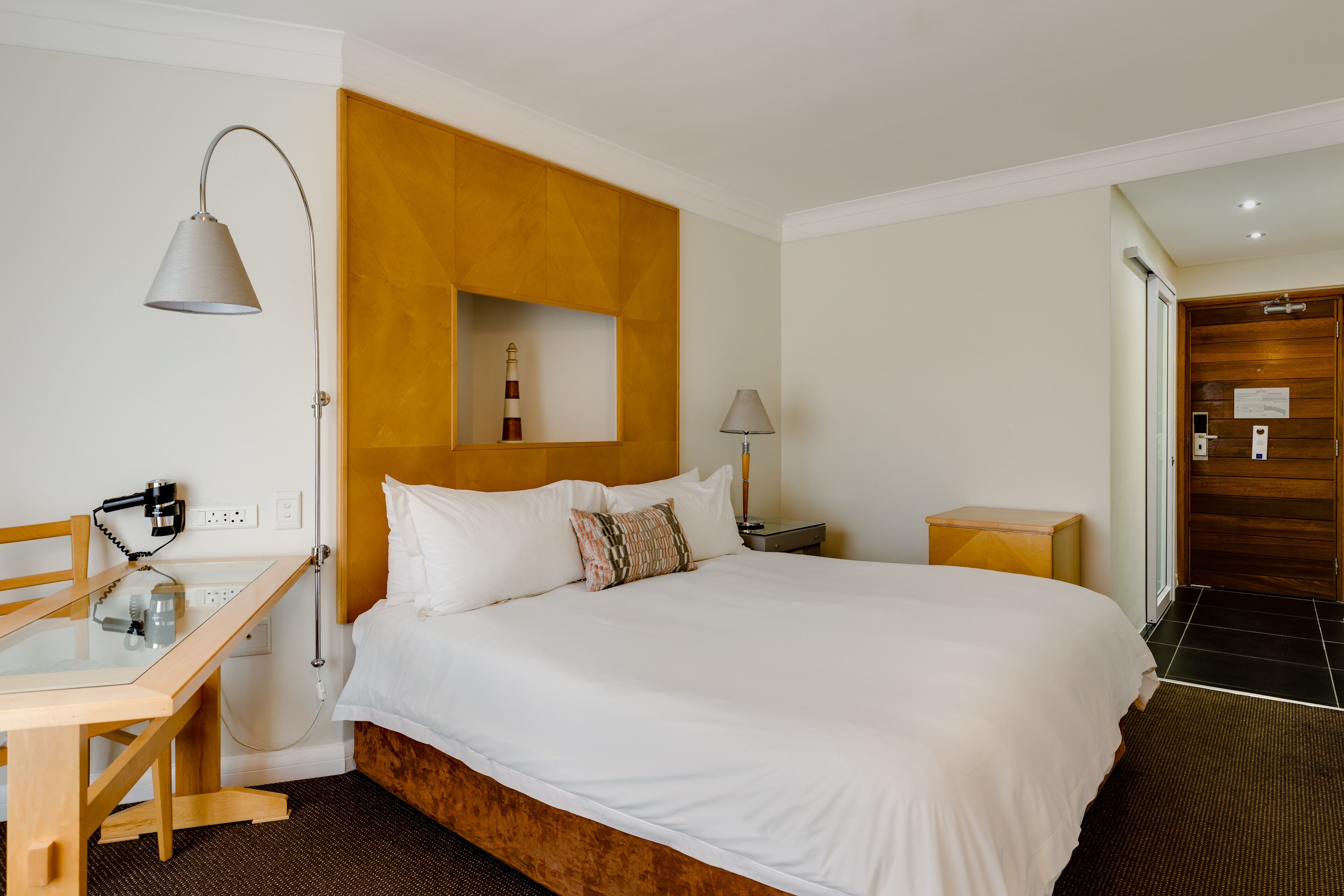A hotel room with a wooden headboard, a bed with white bedding, a desk with a lamp, and a small nightstand.