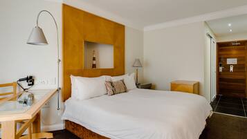 Quarto de hotel com cabeceira de madeira, cama com roupa de cama branca, escrivaninha com candeeiro e mesa de cabeceira pequena.
