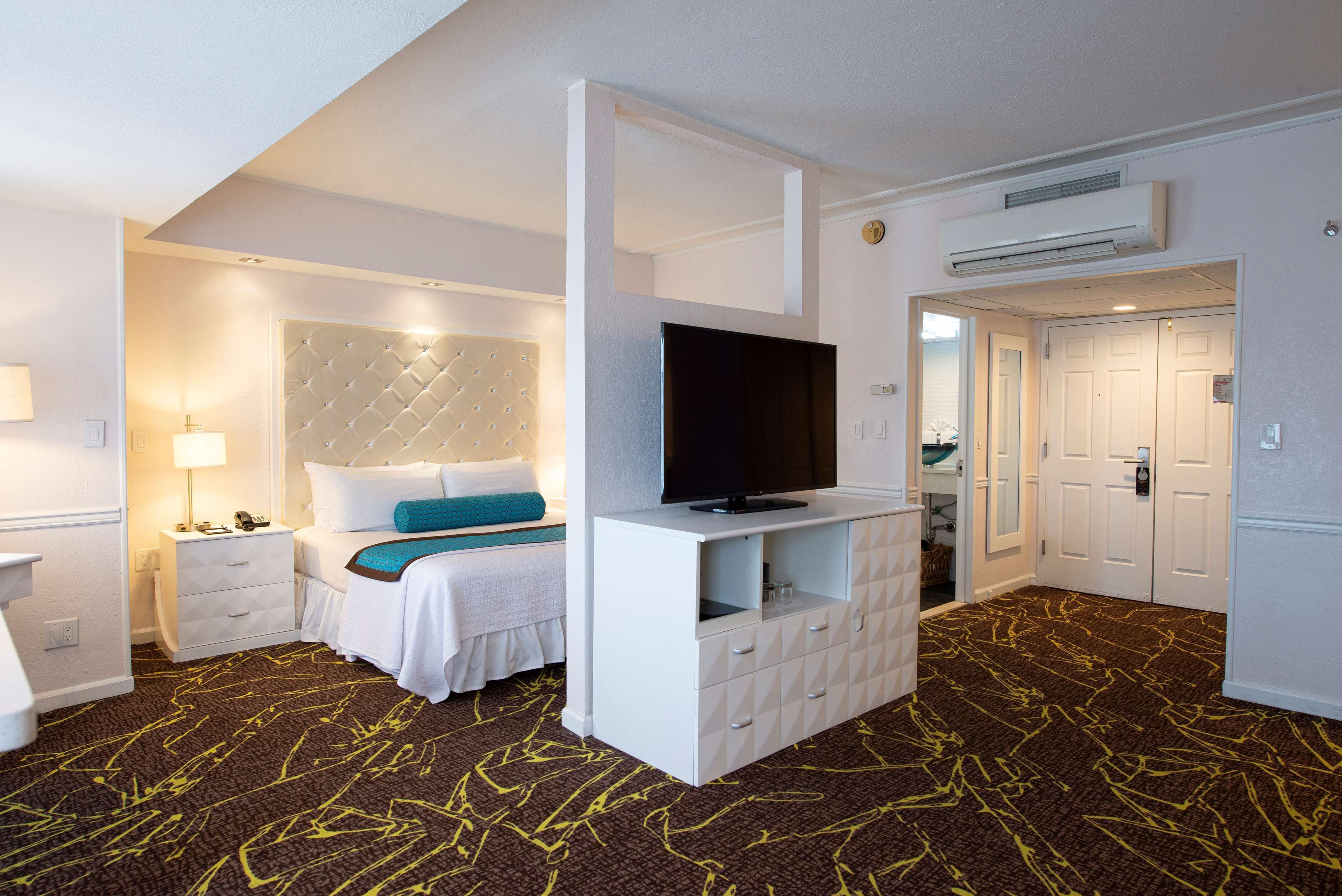 A hotel room with a bed, a television on a stand, a white dresser, and an air conditioning unit on the wall.