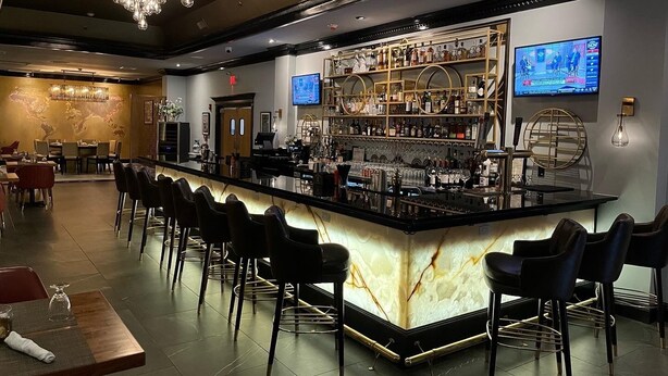A bar area with a marble countertop, multiple bar stools, a world map on the wall, and a television screen displaying a news program. - The Roslyn, Tapestry Collection by Hilton (Roslyn)