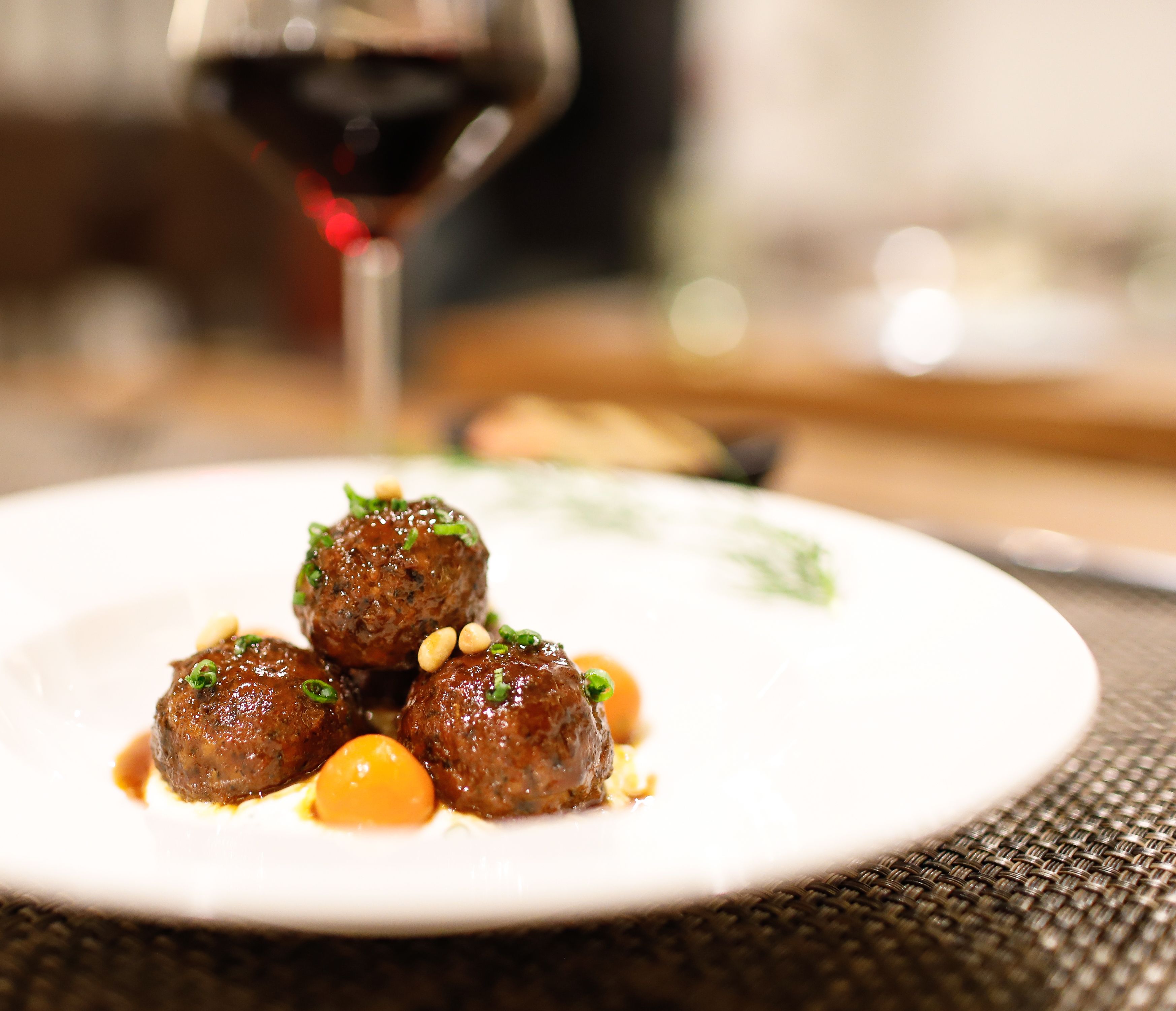 A plate of meatballs with sauce and garnish, a glass of red wine, and a blurred background.