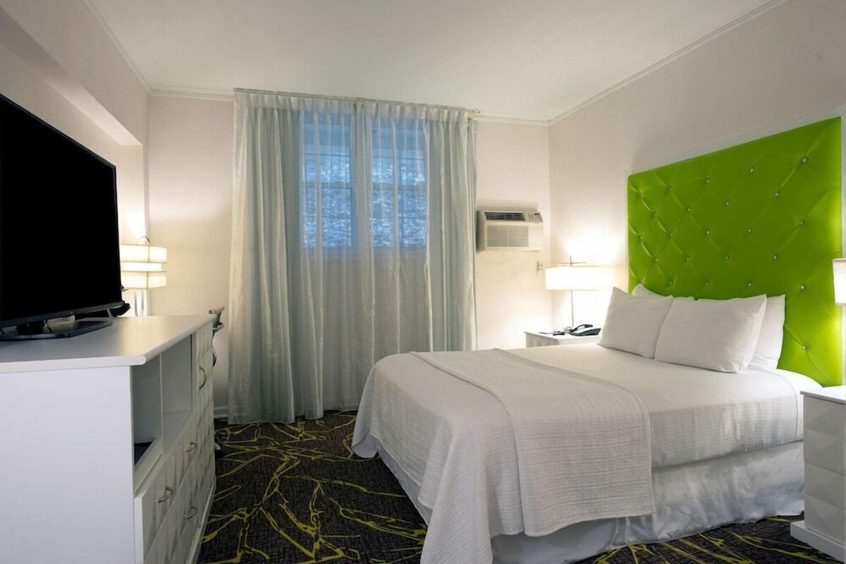 A hotel room with a bed, a television on a dresser, a window with curtains, and a green headboard.