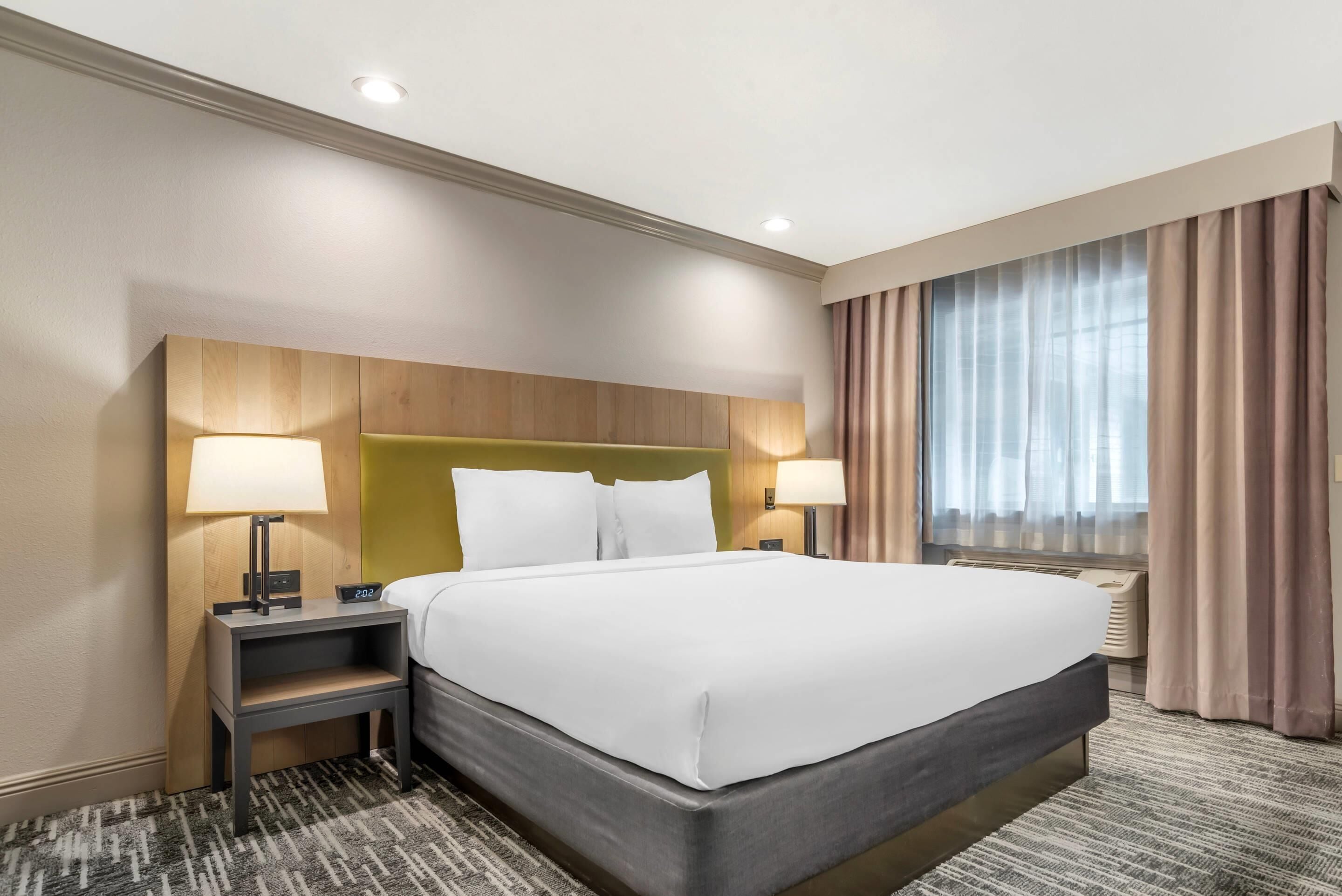 A hotel room with a large bed, two bedside tables with lamps, a headboard, and a window with curtains.