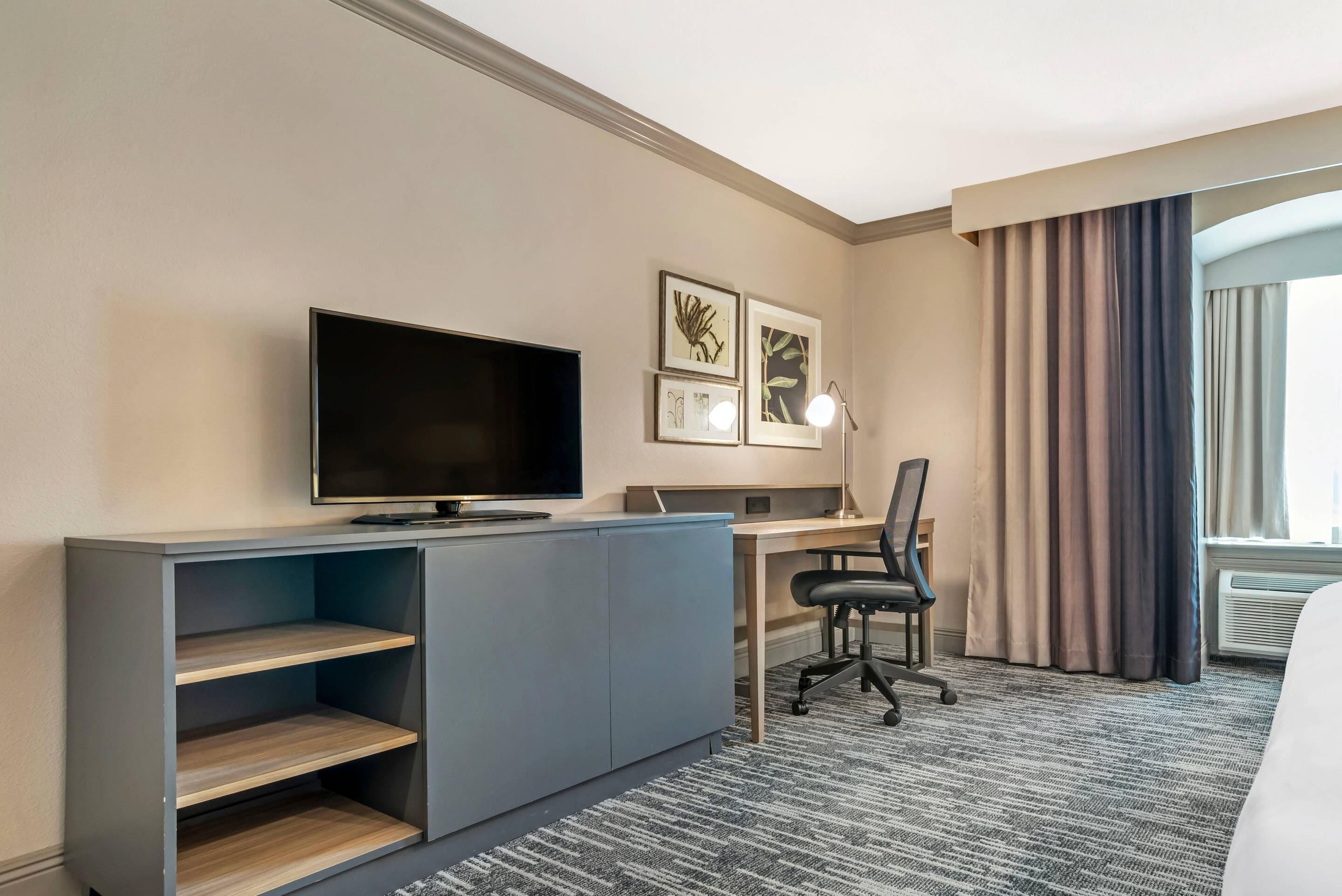 A hotel room with a flat-screen TV, a desk with a chair, a small cabinet, and a window with curtains.