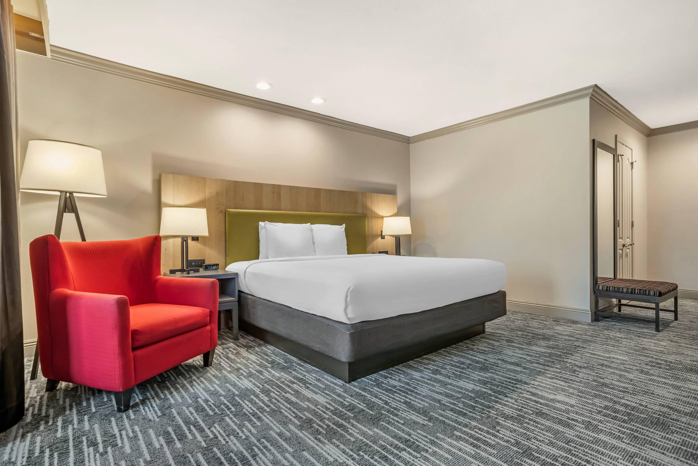 Une chambre d’hôtel avec un lit, un fauteuil rouge, deux tables de chevet avec des lampes, un petit banc et une porte.
