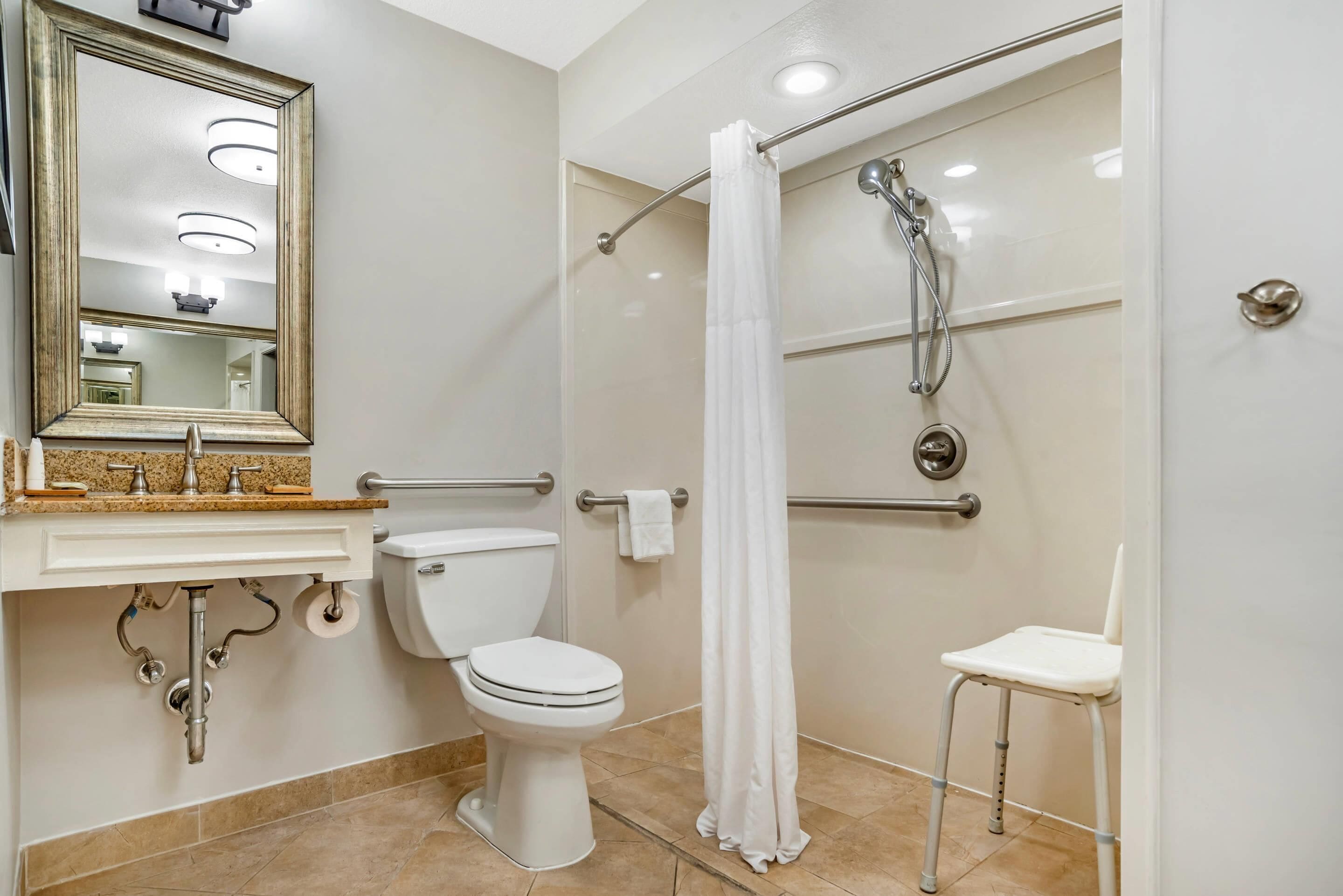 Un baño con inodoro, lavamanos, espejo, ducha y una silla accesible para sillas de ruedas.