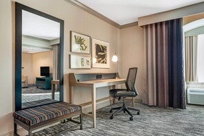 Een hotelkamer met een bureau, stoel, bankje, grote spiegel, kunstwerk en gordijnen.