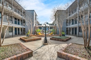 A courtyard with a central light fixture, surrounded by brick planters and trees, with multi-story buildings on either side.