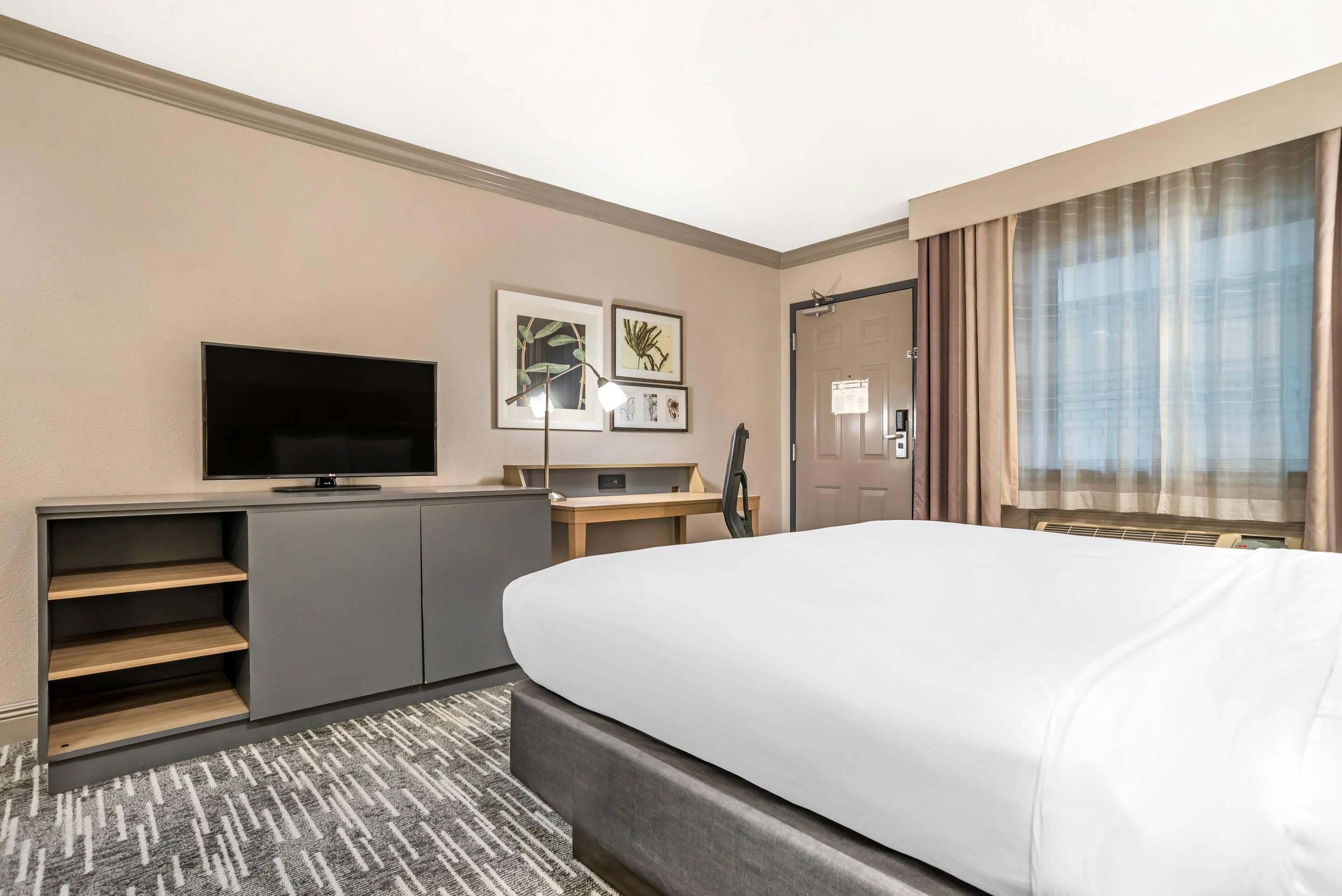 A hotel room with a bed, a TV, a desk with a lamp, and framed pictures on the wall.
