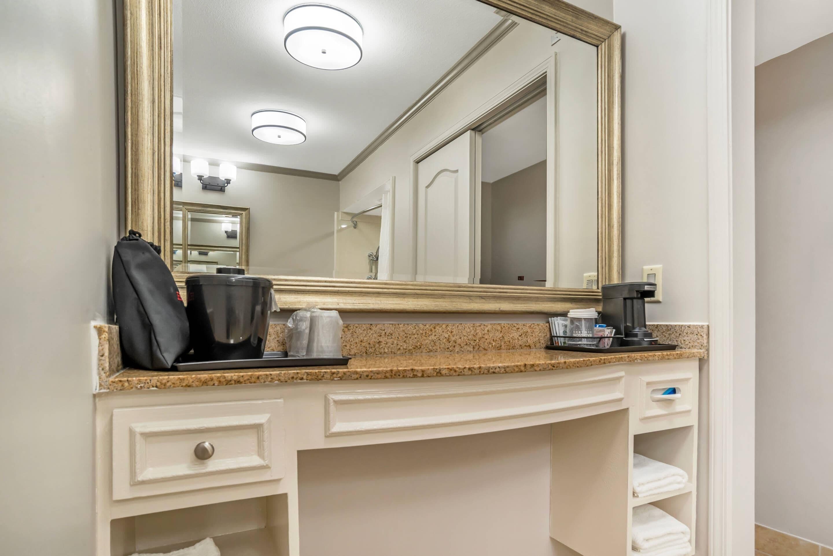 Une salle de bain d’hôtel avec un grand miroir, un plan de travail avec une machine à café et un porte-serviettes.