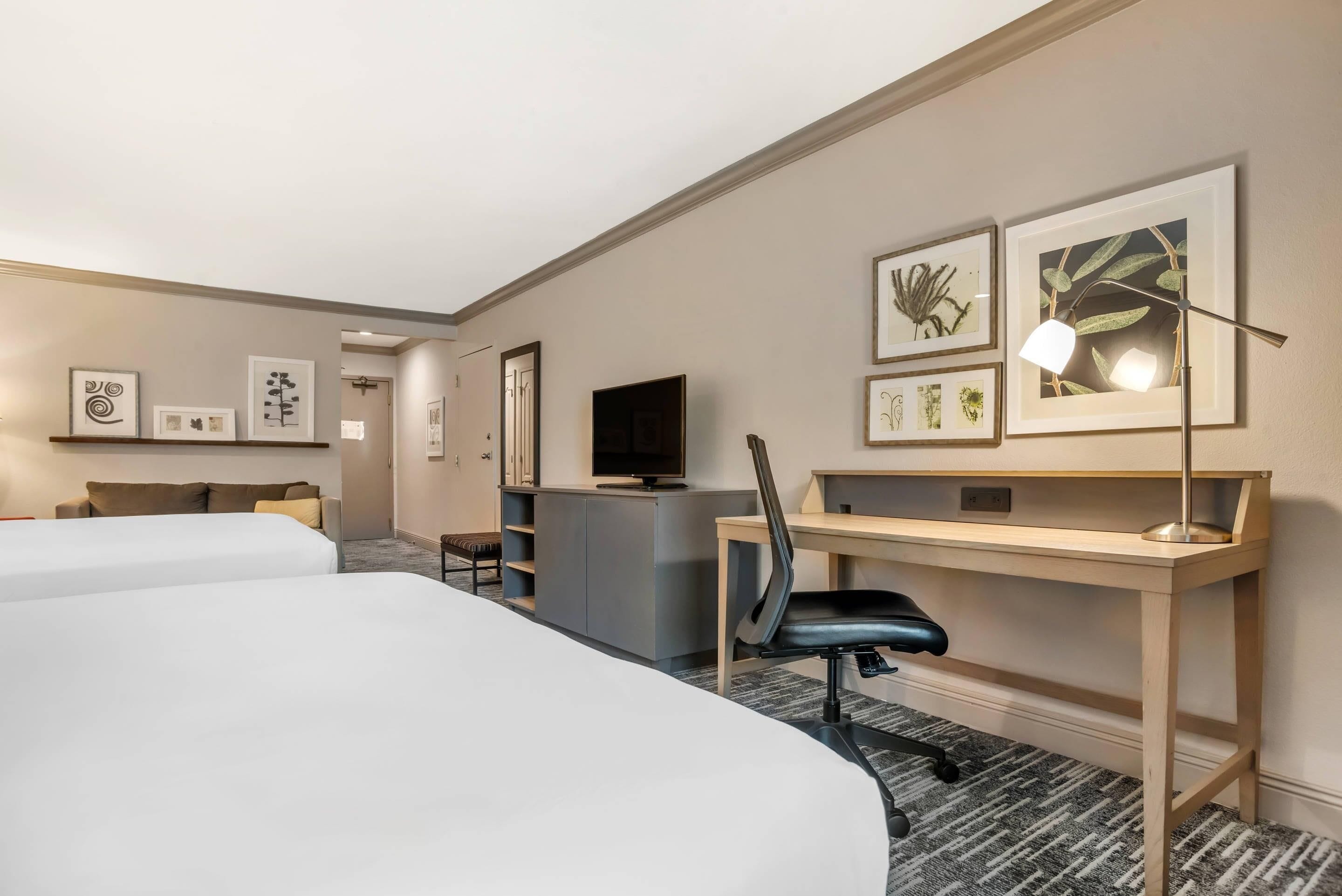 A hotel room with two beds, a desk, a chair, a TV, and framed artwork on the walls.