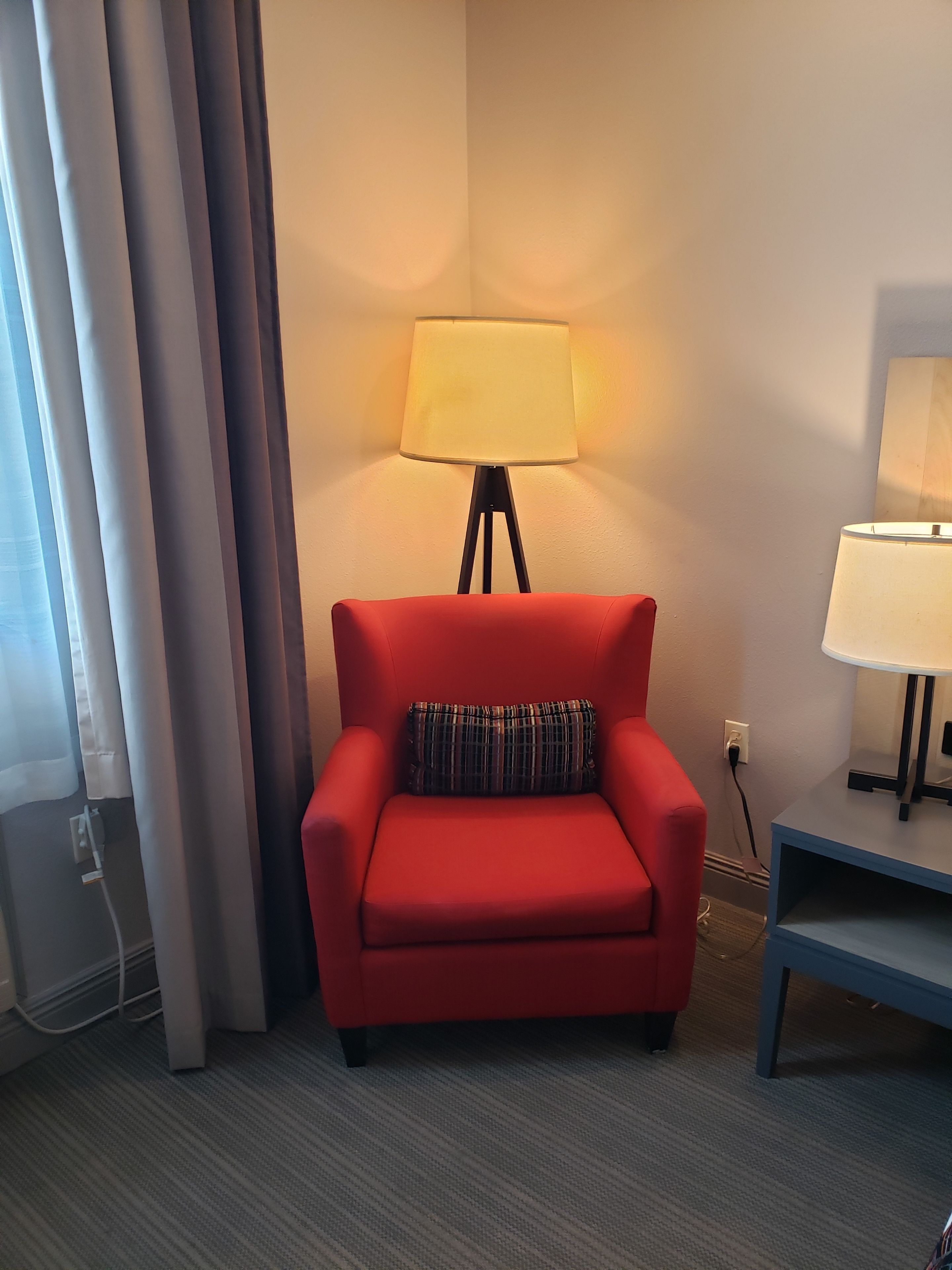 A red armchair with a plaid pillow, a floor lamp, and a small table with a lamp.